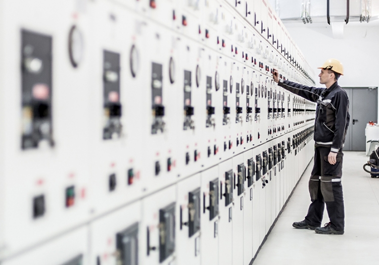 Person in a dark jacket and hard hat standing in front of a control panel with switches and indicators in an industrial setting