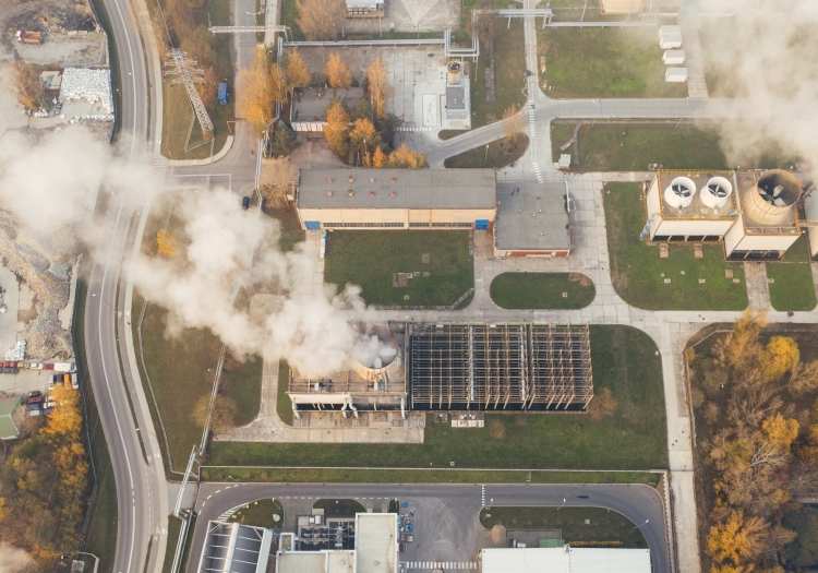 Aerial view of an industrial facility with buildings emitting steam or smoke from stacks, surrounded by roads and autumn-colored trees