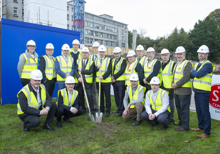 Group of individuals at a construction site wearing safety helmets and high-visibility jackets, gathered around a shovel in the ground for a ground-breaking ceremony