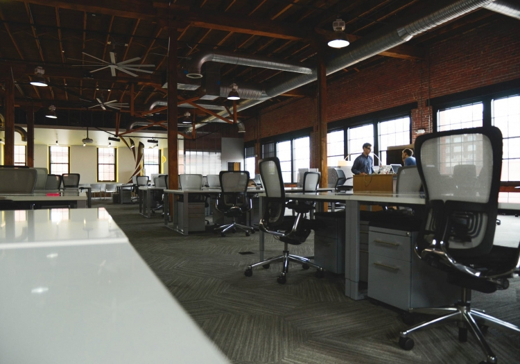 Modern office with desks, computers, ergonomic chairs, exposed wooden beams, brick walls, and large windows. Two individuals converse near a desk in the background