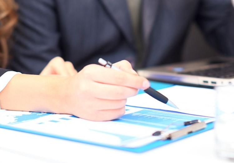 Close-up of a person in business attire holding a pen and resting a hand on a clipboard with a graph or chart. The background includes part of another individual using a laptop, with visible keyboard and screen