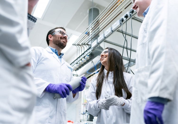 Group of scientists in a laboratory wearing lab coats, gloves, and safety glasses. Two individuals are discussing an object in the center, highlighting research and collaboration