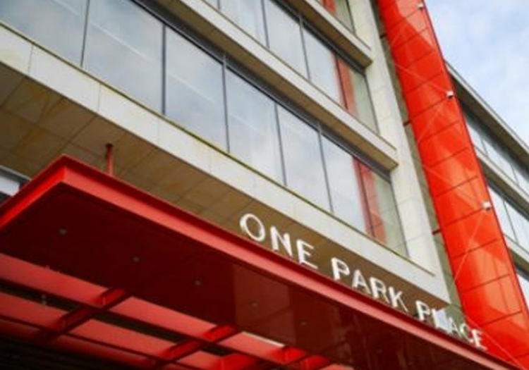 Modern building with several floors, featuring red and grey exterior elements. The red entrance canopy prominently displays “ONE PARK PLACE” in white capital letters. The perspective is from ground level, looking up towards the sky, capturing the building’s façade and the cloudy sky above.