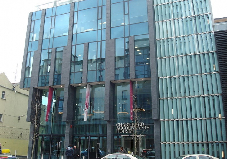 Modern building with a glass facade and vertical metal fins. The entrance features a sign reading “Chartered House” in capital letters, flanked by red banners. The ground floor is pedestrian-friendly, with people near the entrance.