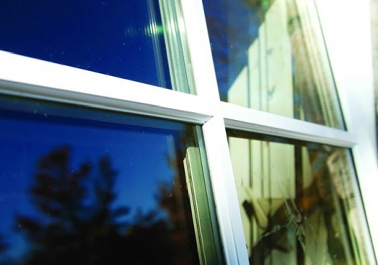 Close-up of a window corner with white frames. The reflection on the glass shows trees and the sky, indicating the window faces a green outdoor area. The angle captures both the reflection and the interior, though details inside are obscured by glare.