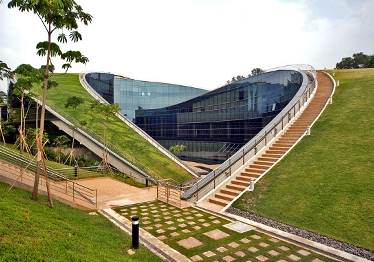 Modern building with a curved glass facade and a grass-covered roof blending into a hillside. The green roof slopes to ground level, with a staircase and accessible ramp leading to the entrance.