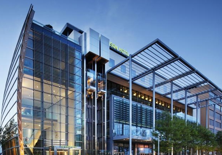 Modern building with a glass facade and multiple levels, featuring steel beams and glass panels for an industrial yet sleek look. Labeled “SONY CENTER” in large, capital letters at the top of the facade. The image is taken at twilight or early evening, with a deep blue sky and visible interior lights. Trees with young leaves in front suggest it is spring or summer.