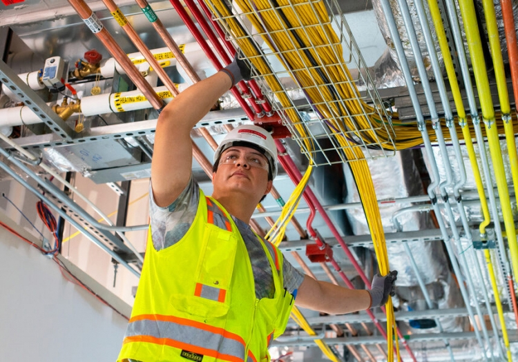 Person in a hard hat and high-visibility vest working on colorful overhead conduits and pipes in an industrial setting