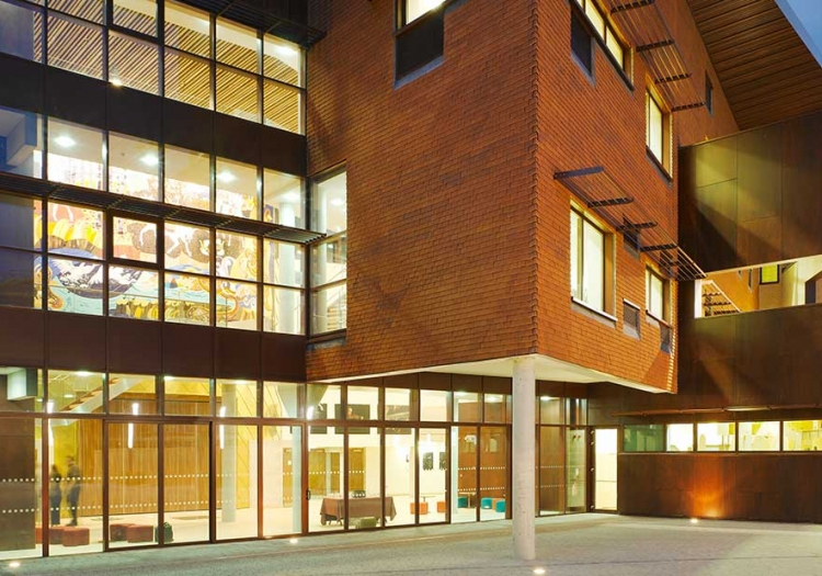 Modern building at dusk with a combination of brickwork and large glass windows. The architecture features interesting angles and overhangs, with interior lights illuminating the rooms, creating a warm and inviting appearance. Benches near the entrance suggest a public or semi-public space designed for multiple users.