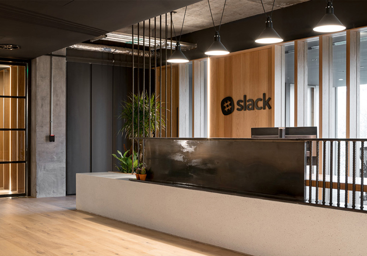 Modern office lobby with a reception desk and the Slack logo on the wall. Well-lit with natural light from glass doors and ceiling fixtures, featuring wood tones and dark colors