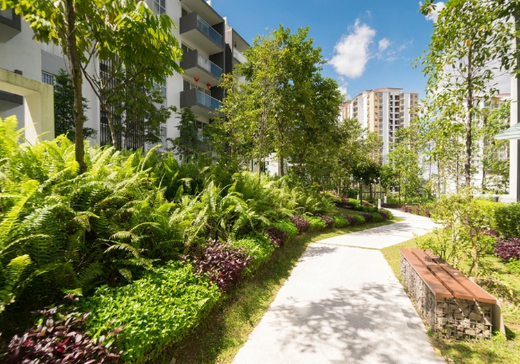 Well-maintained pathway in a residential area with modern apartment buildings. The path is bordered by lush greenery and features a wooden bench. The clear sky and few clouds suggest a pleasant day, highlighting urban landscaping and nature integration.