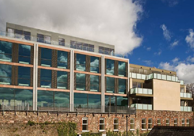 Modern building with a large glass facade and multiple floors, featuring rows of windows reflecting the blue sky and clouds. The contemporary design suggests it is an office or apartment building. In the foreground, an old brick wall contrasts with the new structure, highlighting a blend of old and new in the urban environment.