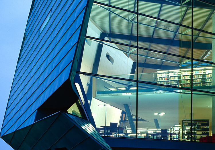 Modern building at twilight with a prominent glass facade. Inside, multiple levels of bookshelves are visible, indicating it might be a library or a building with a significant book collection. The architecture features sharp angles and blue and white lighting, giving it a futuristic appearance.