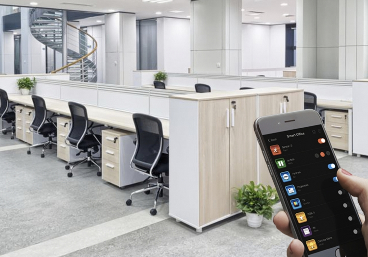 Modern office space with empty workstations featuring desktop computers and black chairs. In the foreground, a hand holds a smartphone displaying application icons. The clean, organized office includes light flooring, white desks, and cabinetry, with a staircase in the background indicating multiple levels. Relevant for discussions on workplace technology, modern office design, or mobile device usage