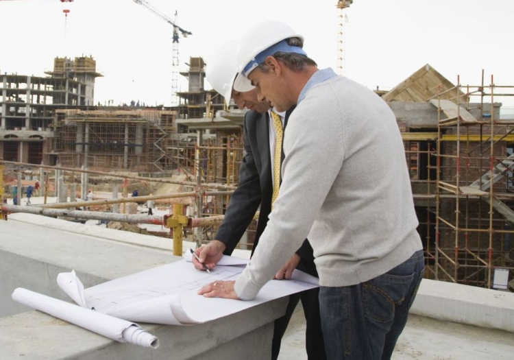 Two individuals at a construction site review a set of architectural plans or blueprints. One person wears a white hard hat, while the other’s face is obscured. The background features an active construction site with structures in various stages of completion and cranes against the sky.