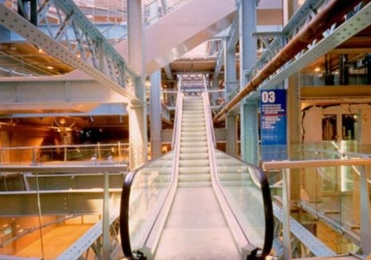 Interior space with modern industrial design featuring a central escalator flanked by metal beams and glass panels. The escalator leads to another floor, indicating part of a larger building, such as a mall or public space. Commercial lighting fixtures and informational signage are visible in the background. The design includes exposed steel girders painted light blue, contrasting with warm wooden elements.