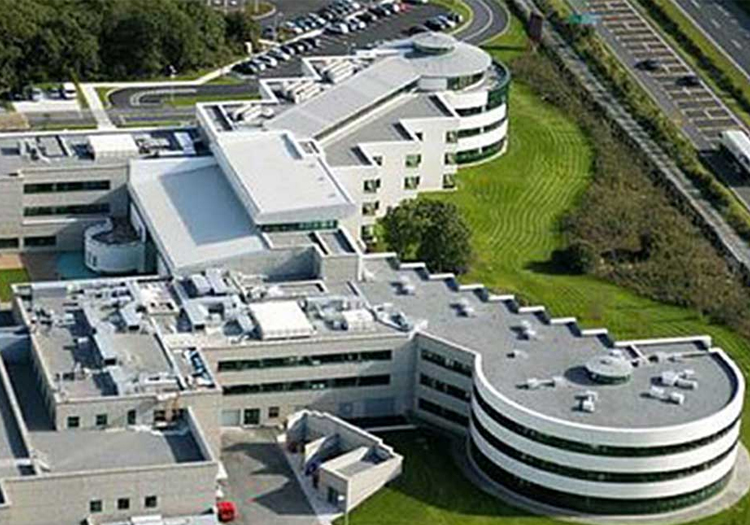 Aerial view of a modern building complex with multiple levels and sections, featuring both curved and straight-edged structures. Surrounded by green lawns, the complex includes a parking lot filled with cars and an adjacent road with marked lanes. The image highlights the design and layout of a contemporary institutional or corporate facility within its environment.