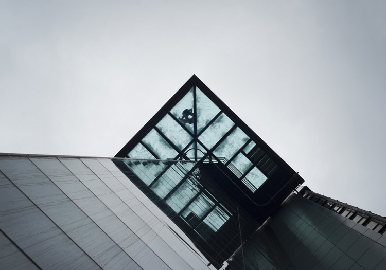 Modern building with an angular design, featuring a protruding glass structure at the top. The overcast sky provides a moody backdrop. The glass section contrasts with the solid parts of the building and reflects the sky and clouds.