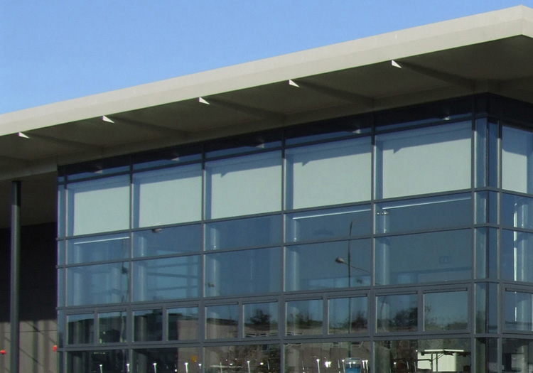 Modern building with a glass facade, flat roof, and multi-level interior. The paved area outside has short metal posts. The visible interior with chairs and tables suggests a public or commercial space