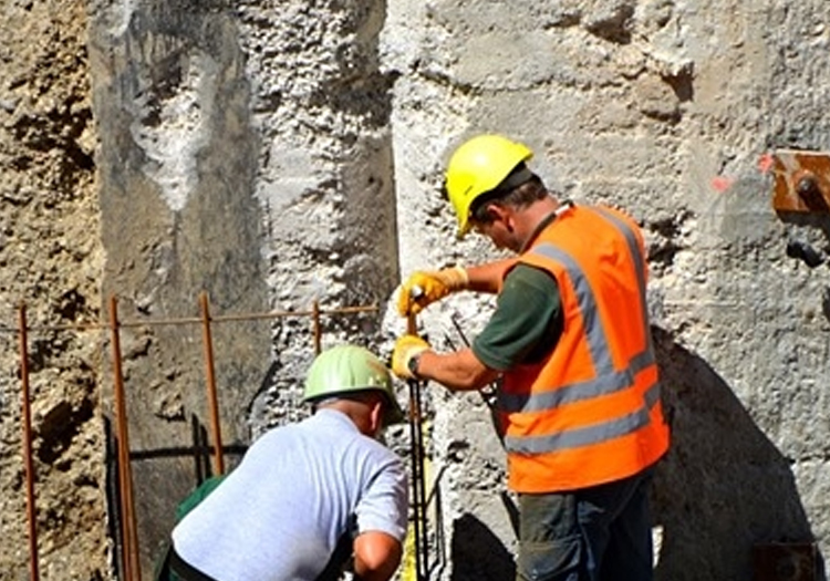 Two construction workers on site: one using a tool on a vertical surface and the other bending over, both wearing safety helmets and high-visibility vests. The background shows a rough concrete wall with exposed rebar, indicating ongoing construction or repairs