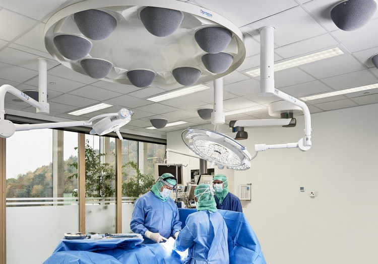 Operating room with medical staff performing a procedure, overhead surgical lights, monitors, and a large window with a view of trees outside