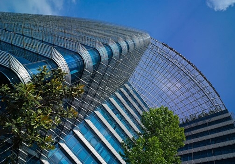 Modern skyscraper with a curved glass façade and visible steel beams. The view is from the ground looking up at the blue sky with clouds, framed by green trees at the base