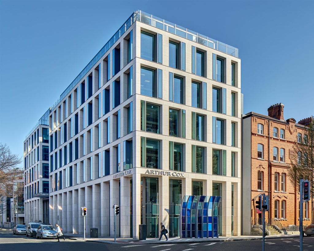 Modern multi-story building on a street corner with a glass and concrete facade. The ground floor has large windows, and 'ARTHUR COX' signage is above the entrance. Blue panels add color to the neutral design. The setting includes a clear sky, leafless trees, and older buildings in the background