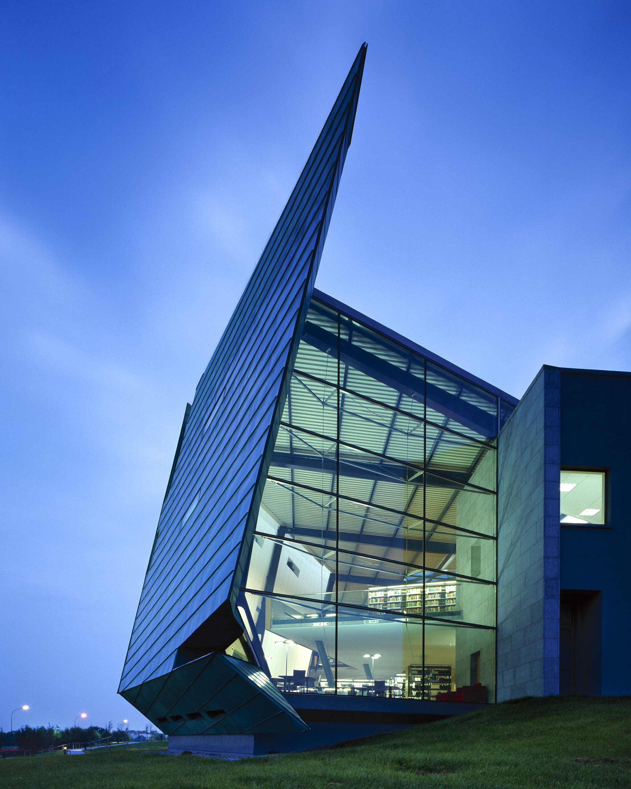 Modern building at twilight with a ship-like angled façade, made of glass and steel, with visible interior lighting through large windows