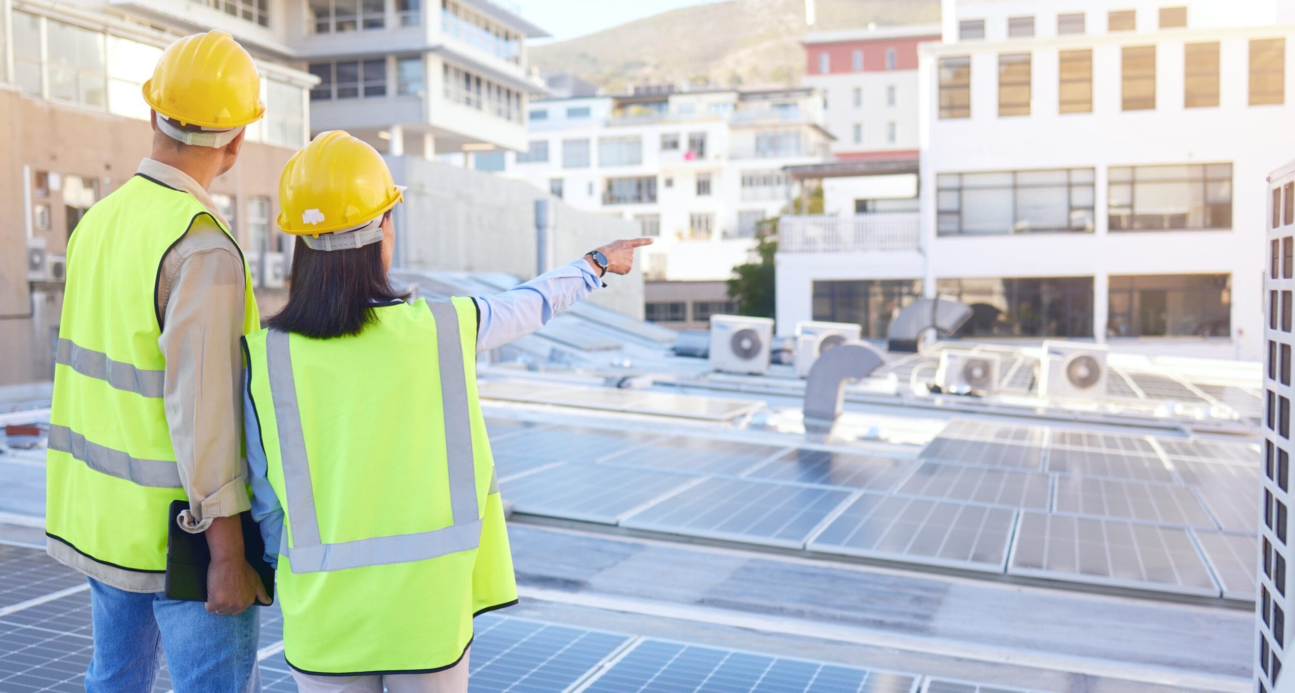 Two individuals in safety helmets and high-visibility vests on a rooftop with solar panels and air conditioning units. One is pointing towards the horizon, likely discussing installation or maintenance