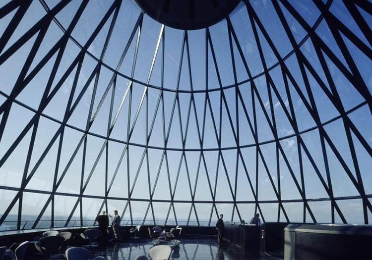 Interior of a modern space featuring a large geometric glass dome with metal beams. Natural light illuminates tables and chairs below, suggesting a public area like a café or gallery. People are seated and standing, enjoying a bright, sunny view through extensive glazing