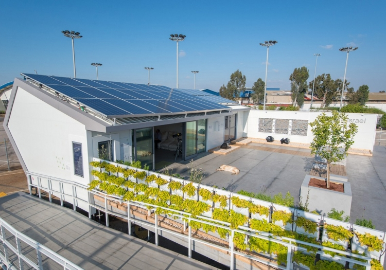 Modern building with a slanted roof covered in solar panels, a balcony with green plants, surrounded by light poles, under a clear blue sky