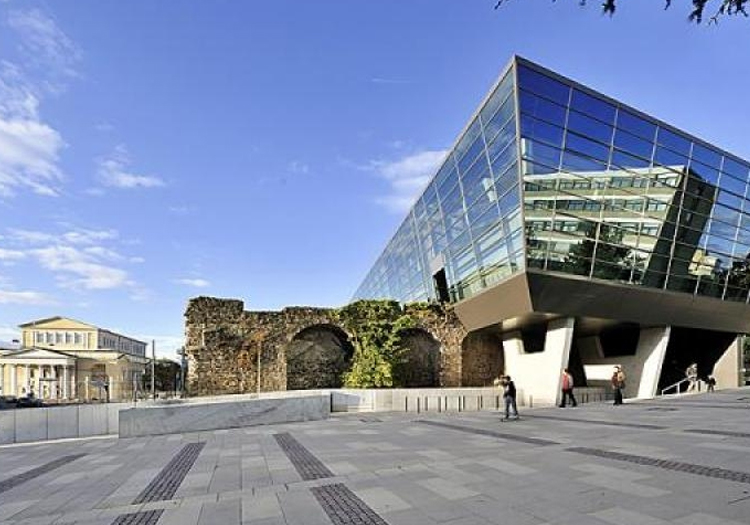 The image features a modern building with a distinctive geometric glass façade set against the backdrop of an ancient stone wall ruin. This juxtaposition of contemporary architecture and historical ruins creates a striking contrast, emphasizing the integration of old and new elements. Several pedestrians walking by add scale and vibrancy to the scene, showcasing how urban spaces can preserve historical elements while embracing modern design.