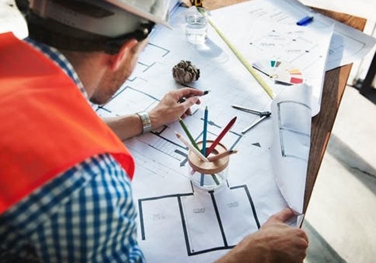 Person in a white hard hat examining architectural plans with a pencil on a table. Nearby items include colored pencils, a pine cone, and a water bottle. The plans feature rectangles and measurements, indicating they are construction blueprints