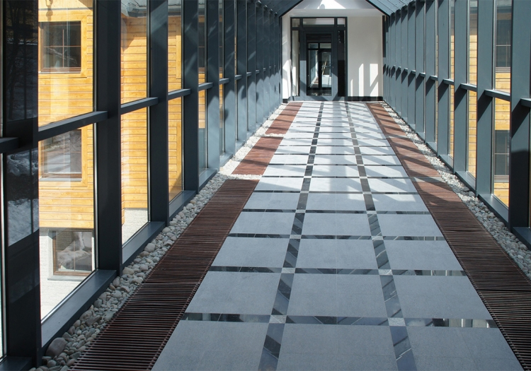 Modern indoor corridor with patterned light and dark tiles, black railings, and large glass windows. A glass door at the end leads to a view of a building with yellow exterior walls