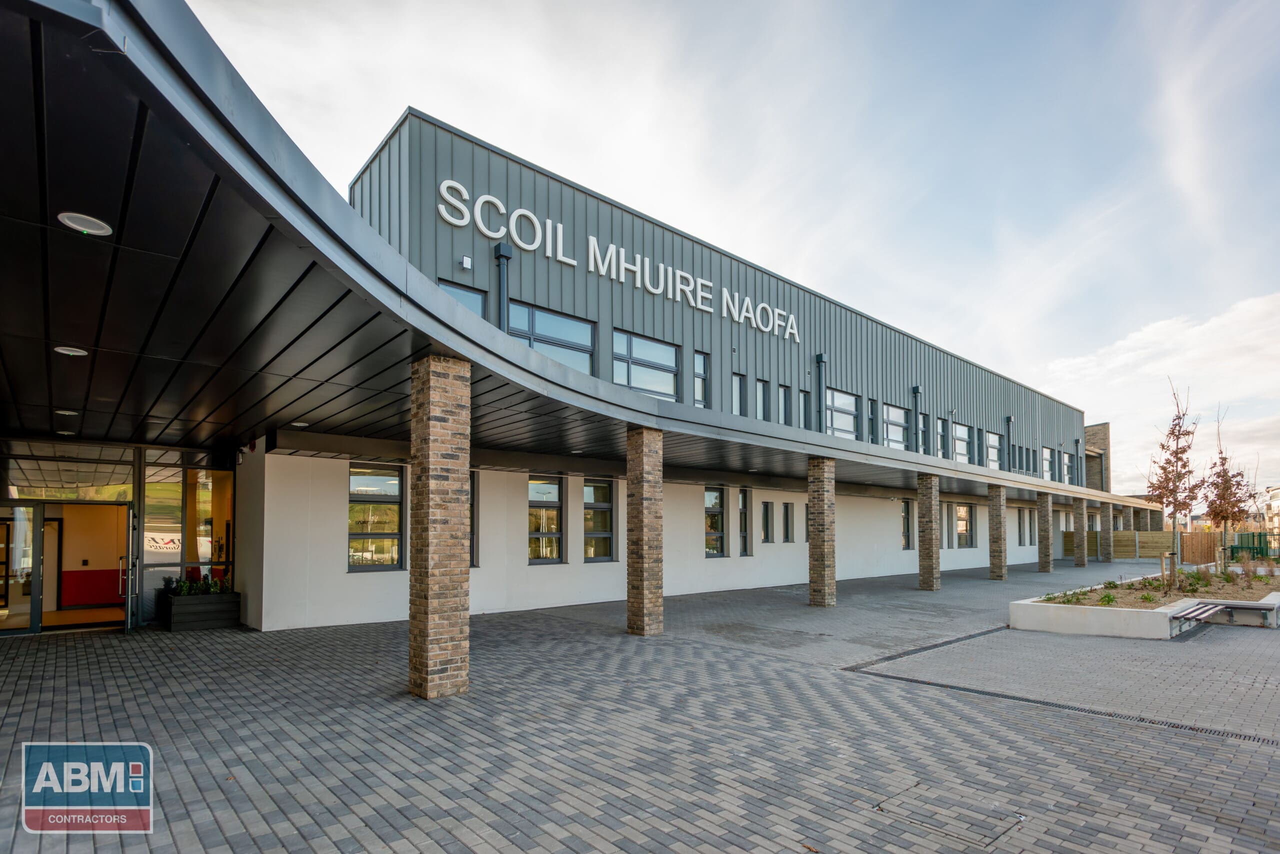 Modern two-story building with a curved roof, featuring a gray facade with wood paneling accents. The ground floor has covered walkways supported by pillars, and the name 'SCOIL MHUIRE NAOFA' is displayed prominently, indicating it is an educational institution. Clear skies suggest fair weather