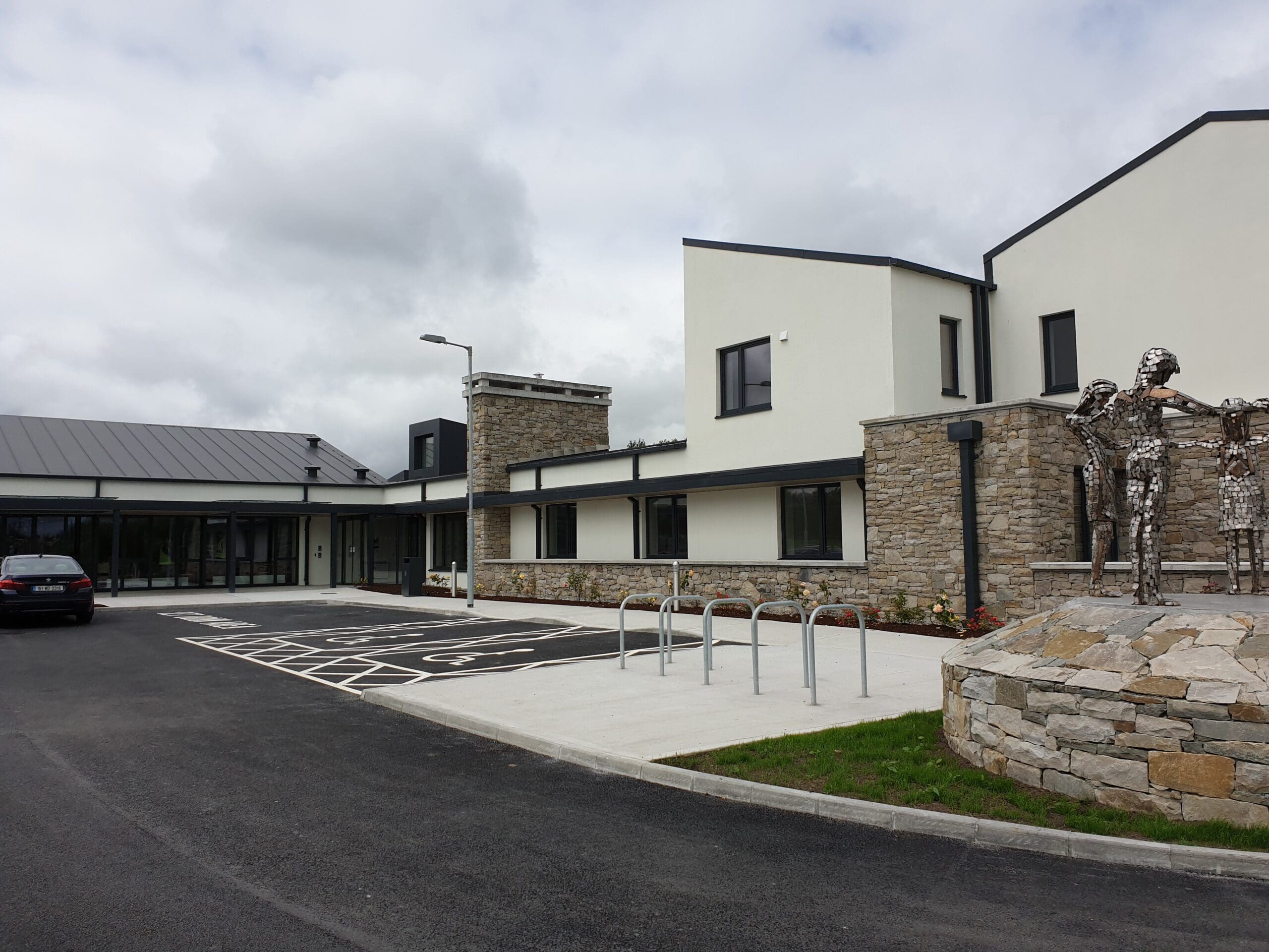 Modern building with white and stone exterior walls. The front features a parking area with marked spaces, including a disabled parking spot. To the right is a metallic sculpture of two human figures