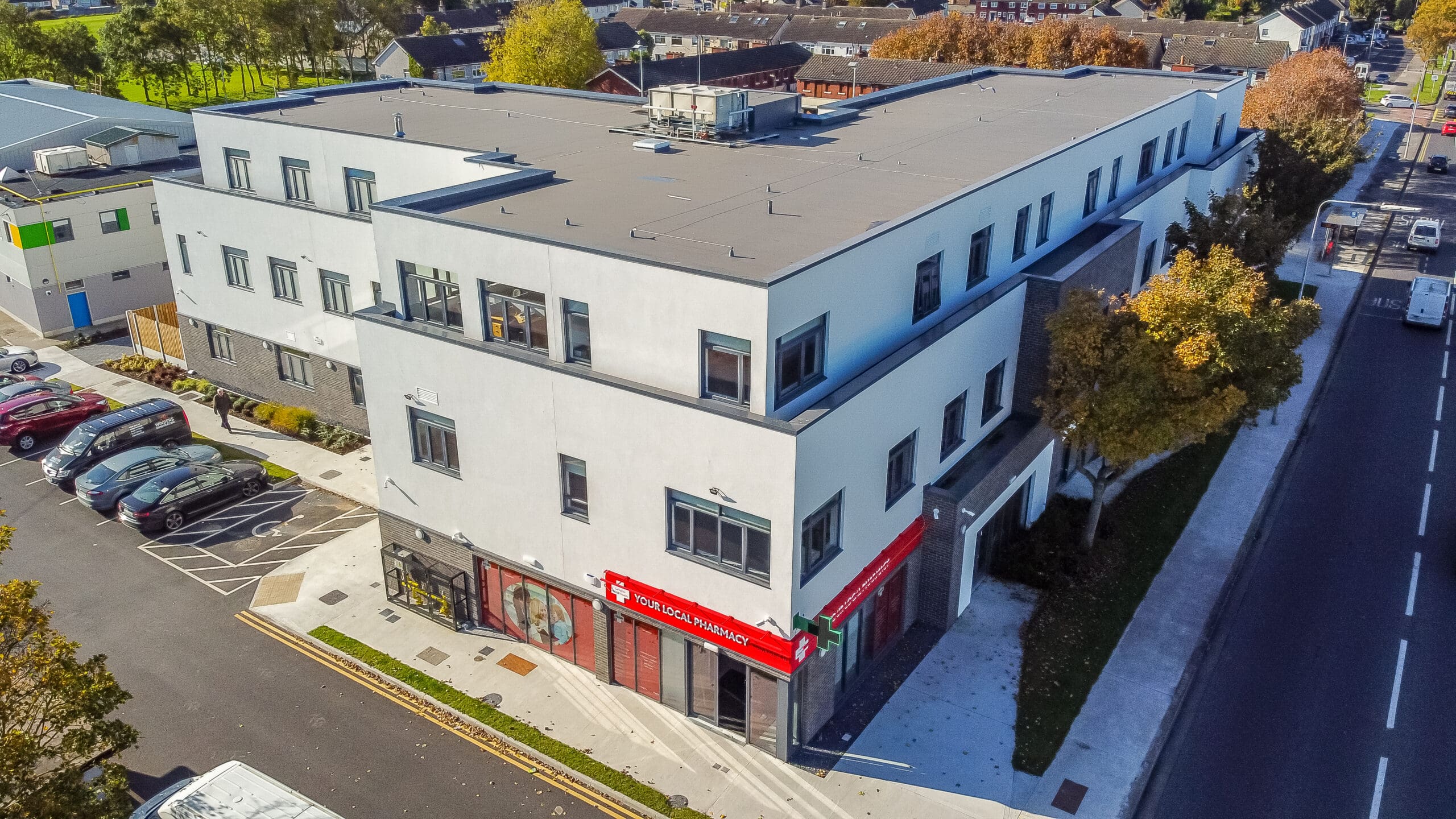 Aerial view of a modern white multi-story building with dark gray accents, surrounded by roads and a parking lot. A red awning marks an entrance or business