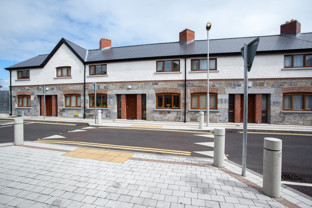 Row of modern terraced houses with brick and plaster façades, individual entrances, and windows on both floors. Street lamps and pedestrian crossing markings are visible on the paved street under a clear sky