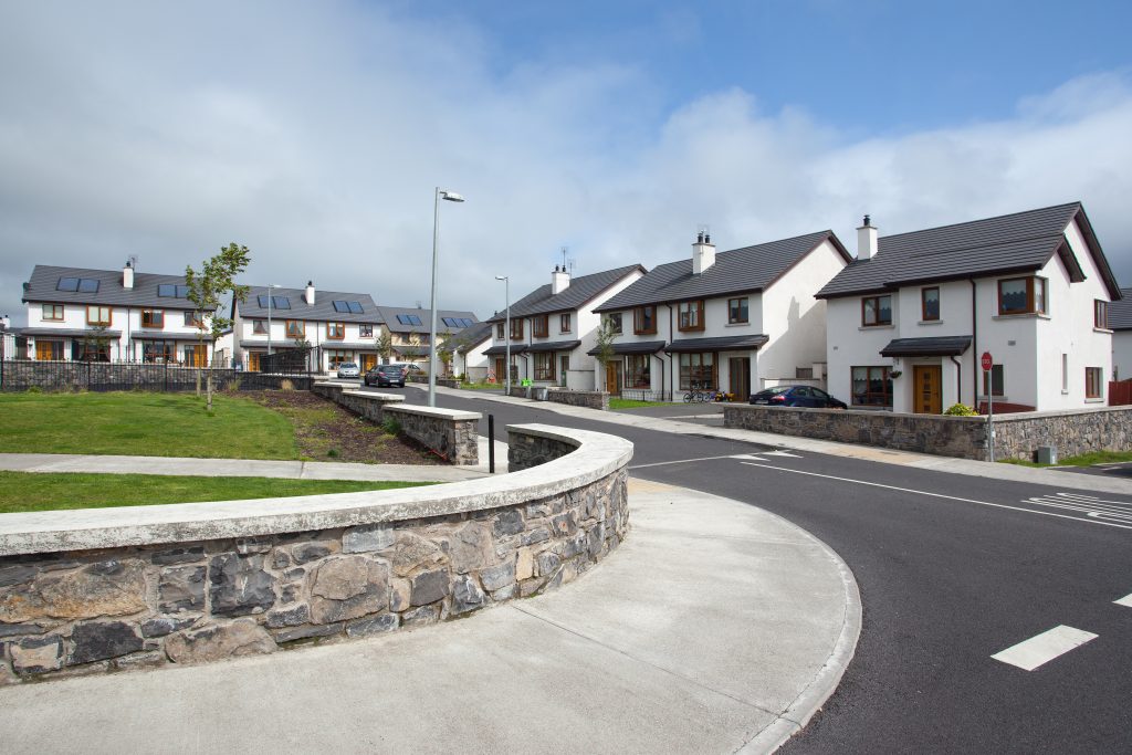 Modern residential area with two-story houses featuring white facades, dark roofs, and dormer windows. Foreground includes a well-maintained lawn with a stone retaining wall and a paved footpath. Partly cloudy sky suggests a pleasant day