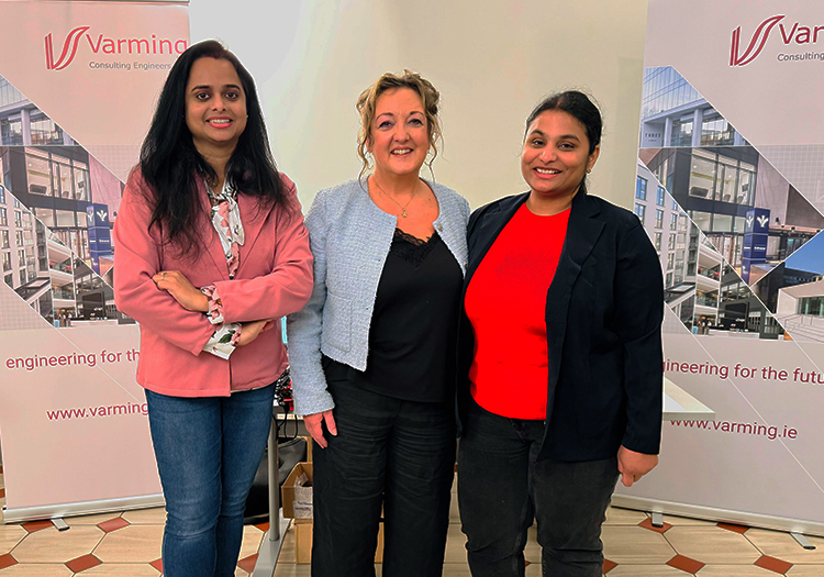 Three women standing in front of Varming Consulting Engineers banners, smiling in a professional setting. The banners display the company logo and tagline "engineering for the future," along with architectural images and the company website URL