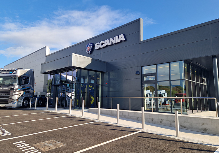 Modern grey building with large glass windows and the Scania logo, featuring a parked Scania truck and visitor parking spaces under a blue sky.